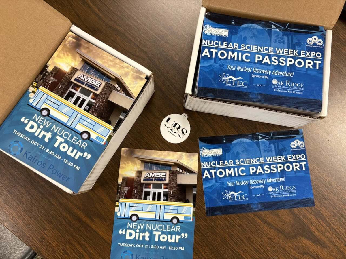 Several printed promotional items for the Nuclear Science Week Expo Atomic Passport are arranged on a wooden tabletop, including blue and white cards, a booklet, and a box. Visible text includes 'NUCLEAR SCIENCE WEEK EXPO ATOMIC PASSPORT,' 'New Nuclear Dirt Tour,' and 'KAFRE Power. Created by United Branding Supply.