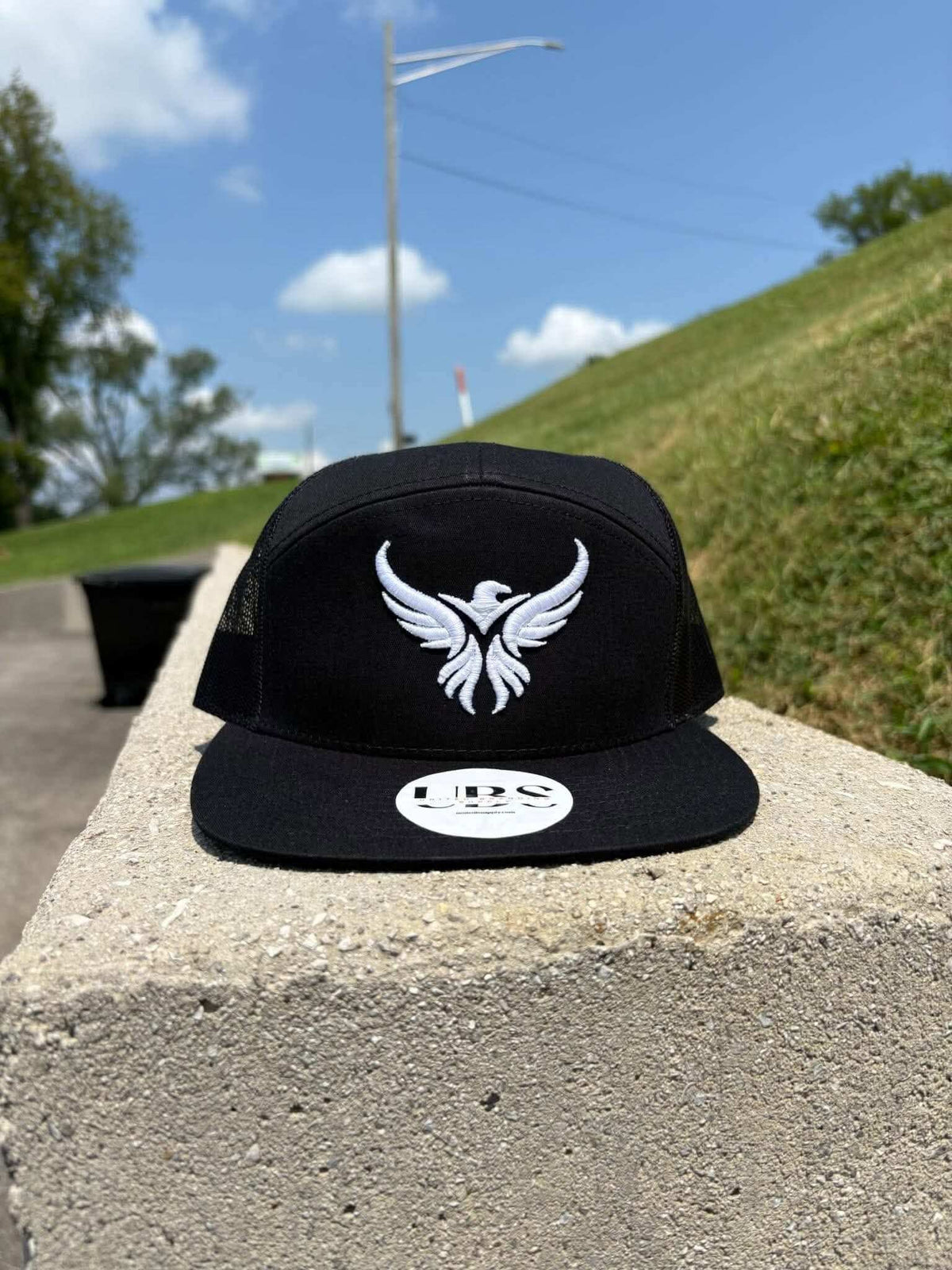 A black trucker-style snapback cap with a white embroidered phoenix logo on the front, resting on a concrete ledge outdoors with a grassy hill and blue sky in the background. Created by United Branding Supply.