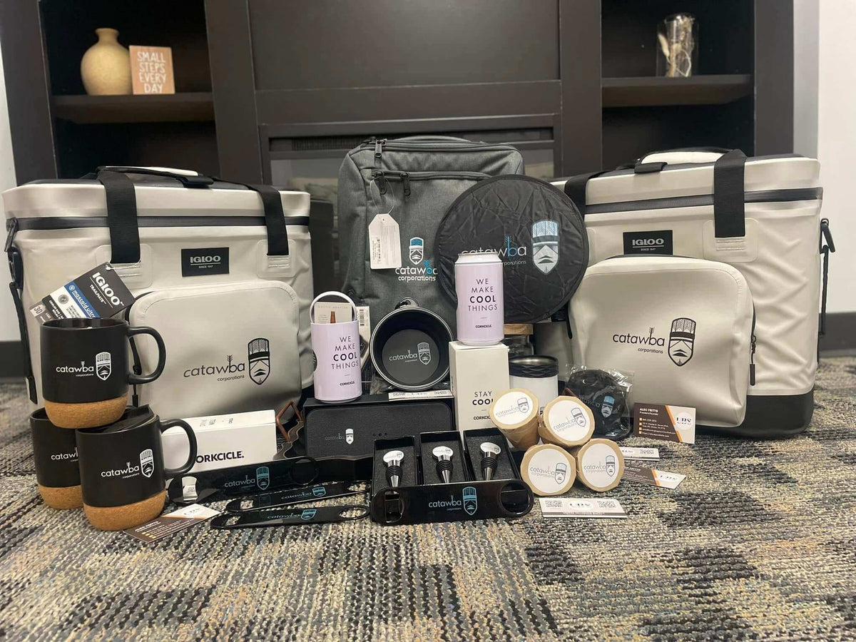 A curated promotional gift set for catawba corporations. Arranged on a patterned carpet in front of a dark wooden cabinet. The set includes two large insulated cooler bags, two black mugs with wood-grain bottoms, a black backpack, a purple insulated bottle, a white coffee maker, several small metal tins, and accessories. Created by United Branding Supply.
