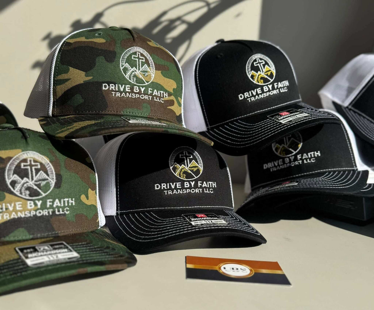 Camo and black trucker hats embroidered with the Drive By Faith Transport LLC logo, by United Branding Supply.