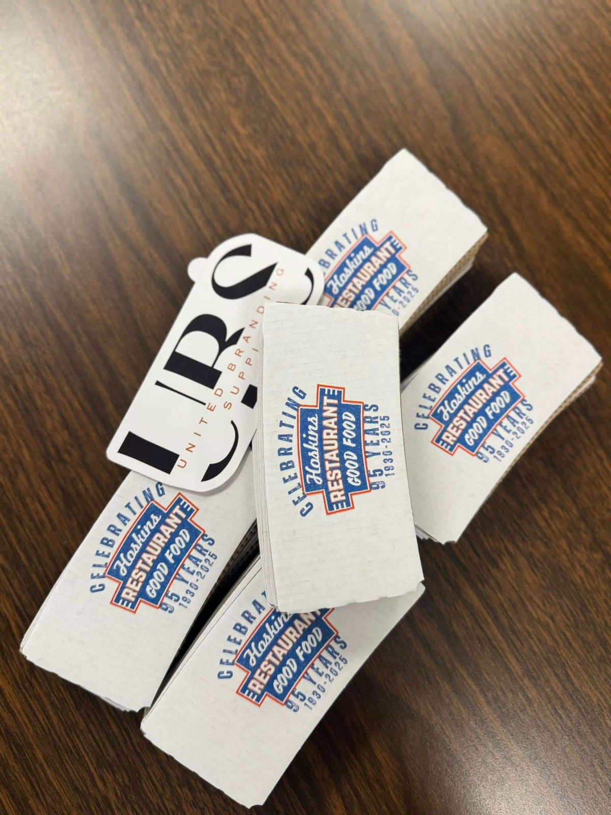 A pile of white cardboard coffee sleeves printed with a restaurant logo that reads 'Celebrating Hoskins Restaurant Good Food. 95 Years. 1930-2025' and a UBS logo sticker laying beside them. Created by United Branding Supply.