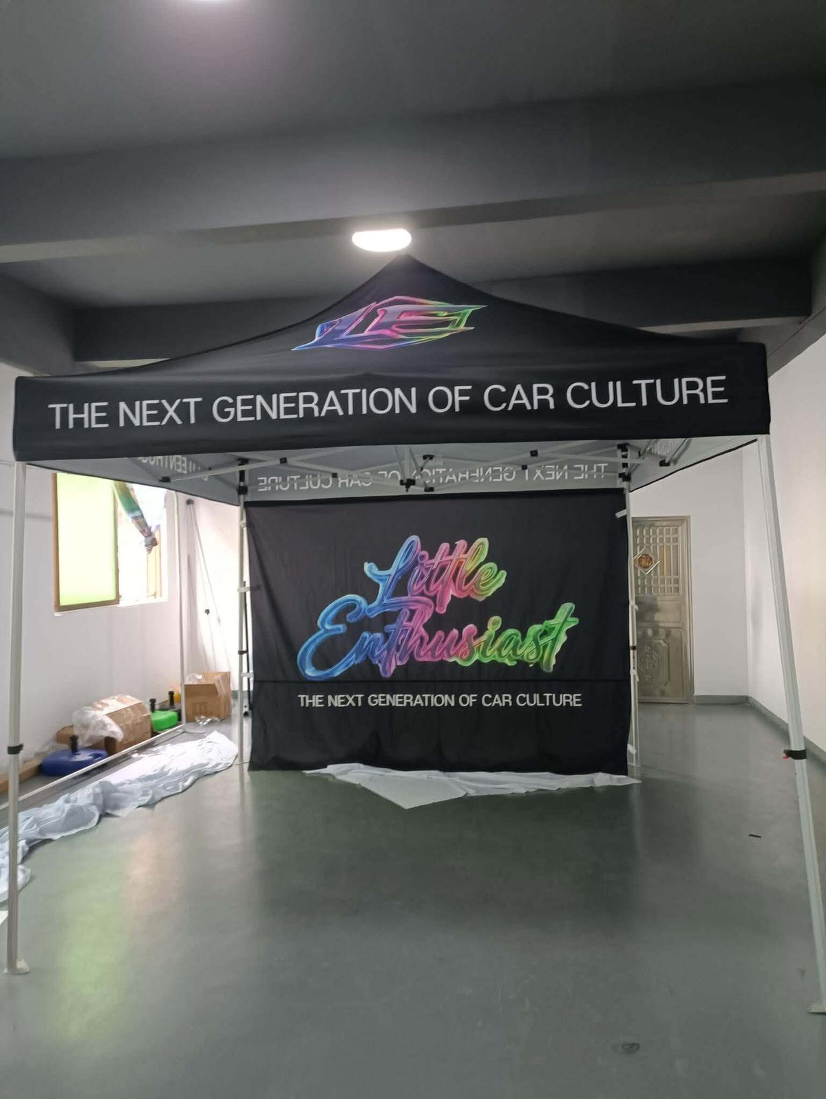A black pop-up canopy tent with custom printed graphics and banners, set up inside a warehouse or studio space. The canopy features the text 'THE NEXT GENERATION OF CAR CULTURE' and a colorful 'Little Enthusiast' logo on the front banner. Created by United Branding Supply.