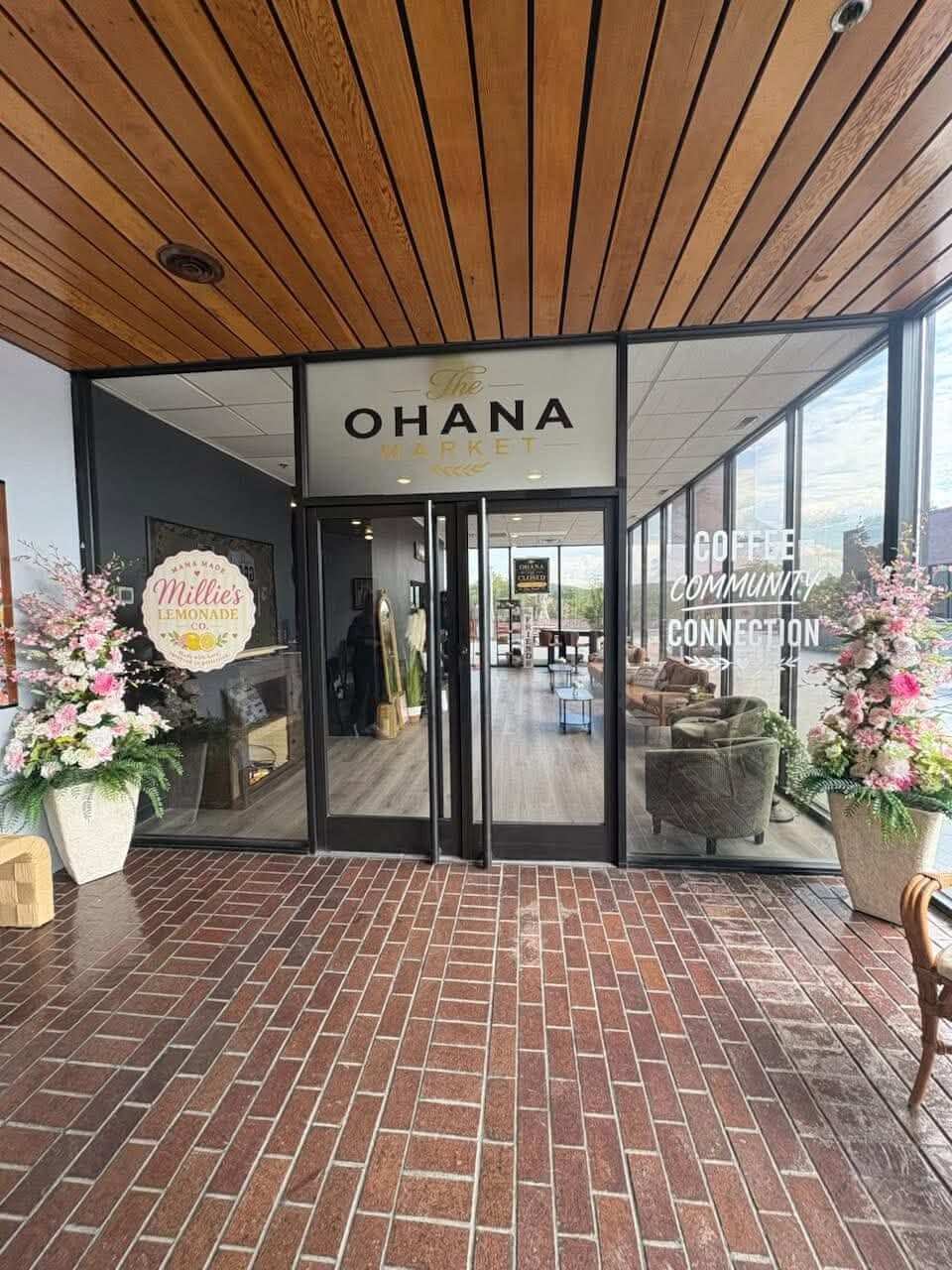 Entrance to The Ohana Market featuring glass double doors, a wood-paneled ceiling, brick flooring, and large floral arrangements on both sides. Window decals by United Branding Supply.