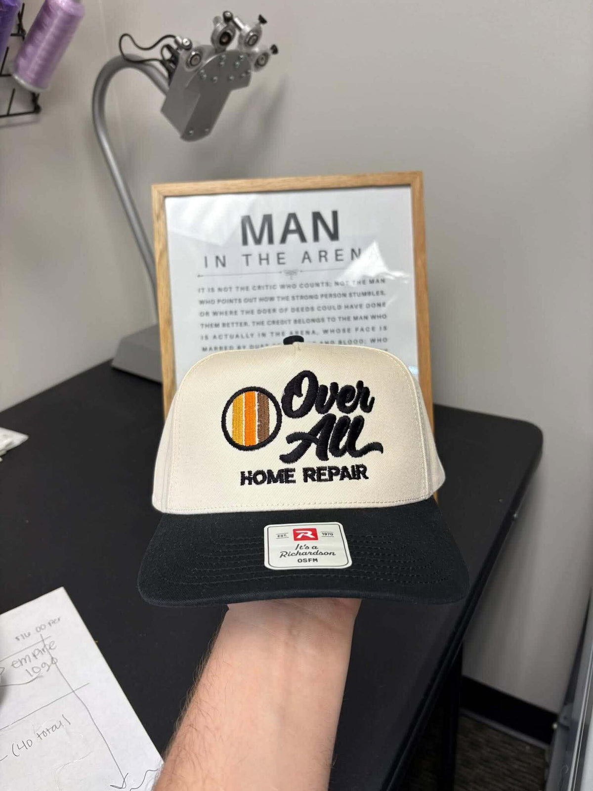 A hand holds a black and beige baseball cap. The cap features a black brim, a beige front panel with 'Over All HOME REPAIR' in black script and block lettering, and a circular orange and black graphic on the left. Created by United Branding Supply.