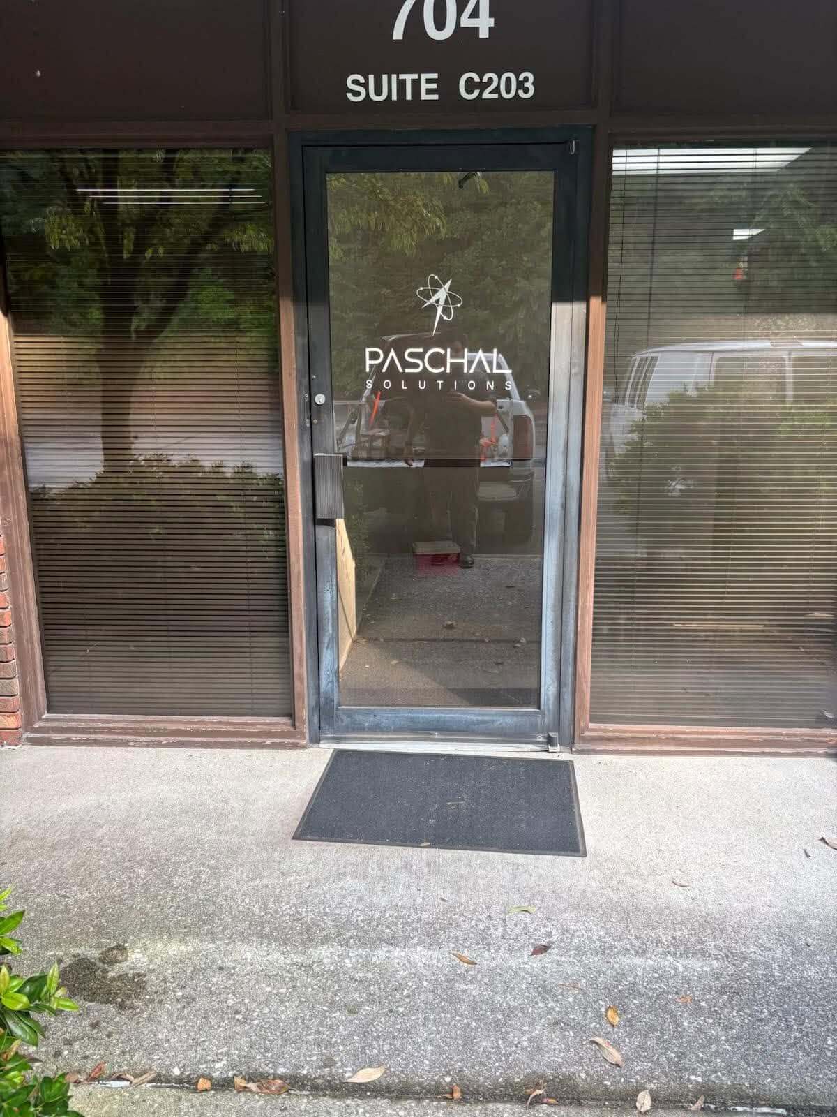 A glass entrance door with the text 'PASCHAL SOLUTIONS' and a logo, set in a storefront. The door is flanked by two windowed sections with closed blinds, and a black doormat sits on the concrete threshold. Window decal created by United Branding Supply.