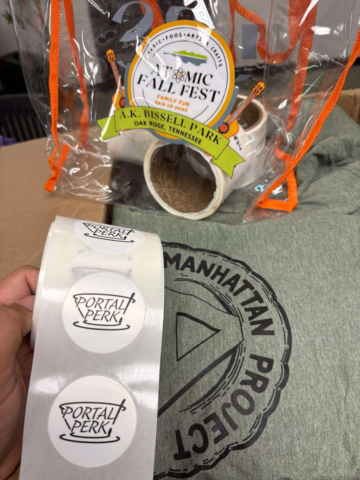A roll of custom Portal Perk stickers, and a Manhattan Project graphic t-shirt, by United Branding Supply