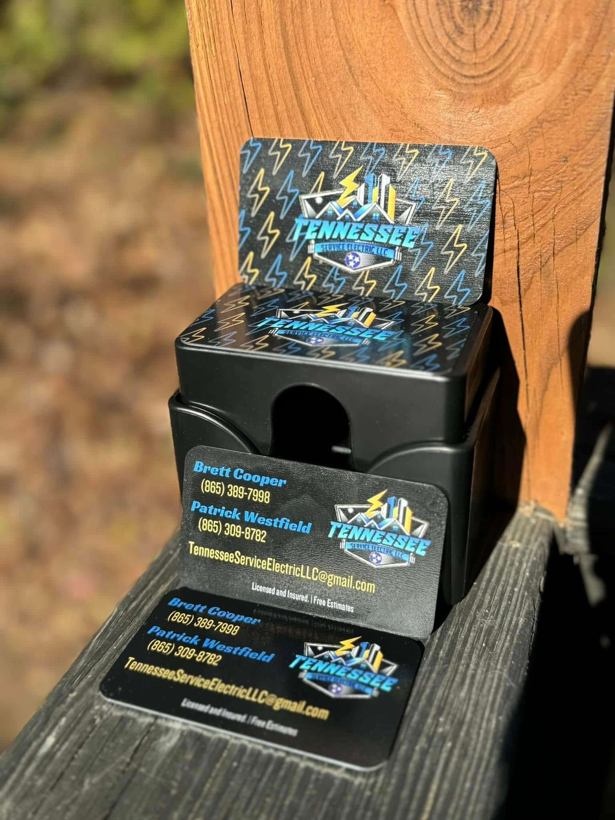A black plastic business card holder displaying Tennessee Service Electric business cards, set against a wooden post and outdoor background. Created by United Branding Supply.