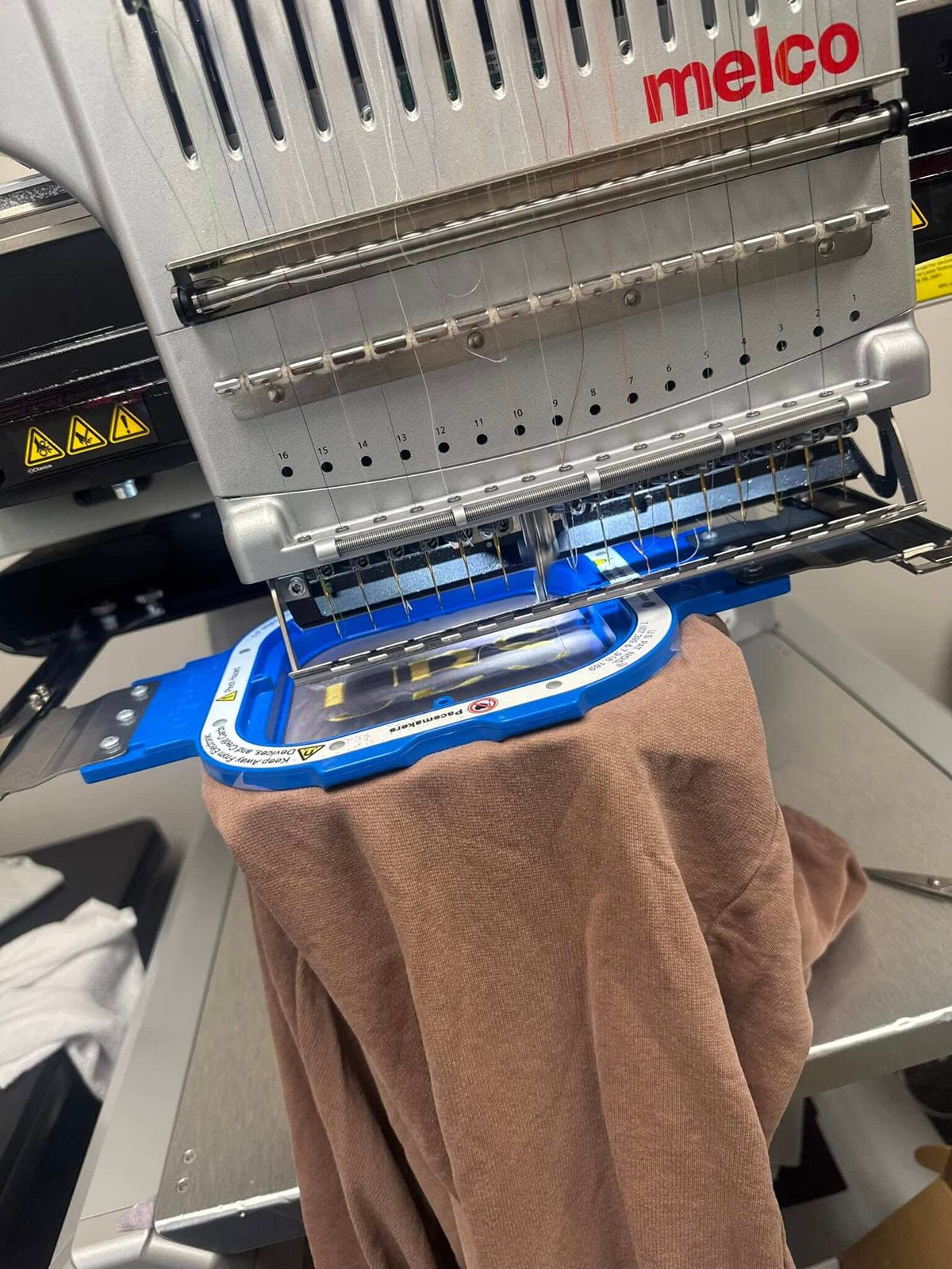 A Melco industrial embroidery machine with a blue hoop holding a brown fabric garment, photographed in a workshop setting. Work in progress at the United Branding Supply Office.